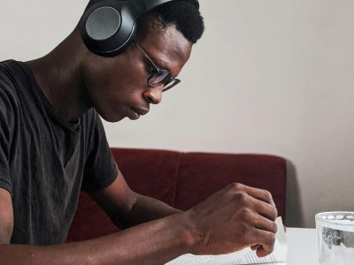 An adult man intensely studying with headphones, pen, and notebook at a desk indoors.