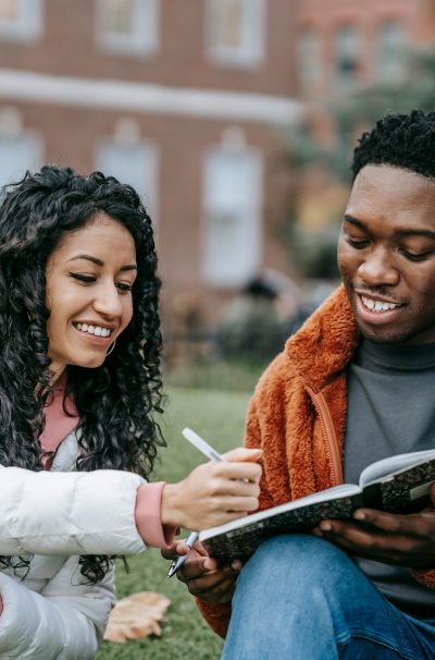 Happy college students studying together on a campus lawn, sharing ideas and writing notes.