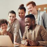 A group of diverse young professionals happily collaborating around a laptop indoors.